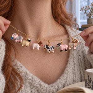 Puede incluir: Un collar de cadena dorada con seis dijes de ovejas en miniatura. Cada dije está coloreado y detallado de forma única, incluyendo ovejas rosas, blancas, negras con manchas blancas y marrones, algunas sosteniendo bayas o flores en la boca.