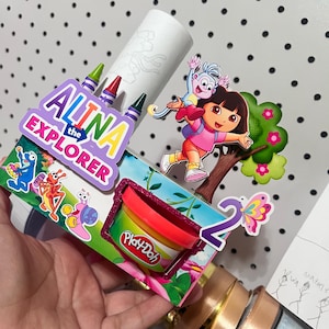 Könnte beinhalten: Ein personalisierter Party-Geschenkehalter mit einem bunten Ausschnitt von Dora und Boots, einem Play-Doh-Behälter und dem Text ALINA the EXPLORER. Das Design umfasst eine Nummer zwei, einen Schmetterling und kleine Buntstifte in einer weißen Röhre.