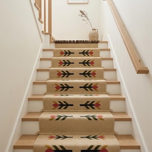 May include: A beige stair runner with a repeating pattern of stylised pink, dark green, and mustard yellow floral motifs. The runner is secured to wooden stairs with metallic gold rods and features dark brown fringe at the top and bottom ends.