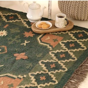 Könnte beinhalten: Ein dunkelgrüner Teppich mit einem sich wiederholenden geometrischen Muster in Beige- und Cremetönen, der mit einer Fransenborte versehen ist. Eine weiße Keramik-Teekanne und eine Tasse mit einem dunklen Getränk stehen auf einem Holztablett mit Keksen.