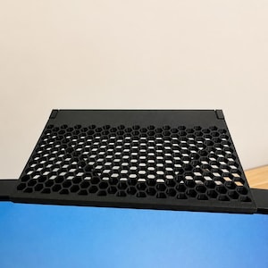 Könnte beinhalten: Ein schwarzes Monitor-Ablagefach mit einem geometrischen Wabenmuster auf der Oberfläche. Das rechteckige Regal ist so konzipiert, dass es an der Oberkante eines Computerbildschirms befestigt werden kann und eine flache, stabile Ablagefläche bietet.