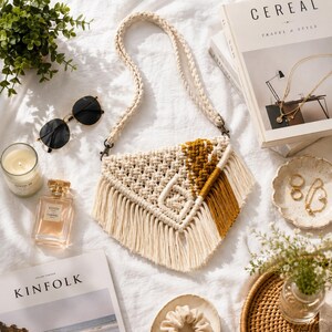 May include: A triangular macrame crossbody bag featuring off-white and mustard yellow cotton cord knots with long fringe trim. The bag includes a braided shoulder strap attached with metal clasps, showcasing a textured geometric pattern.