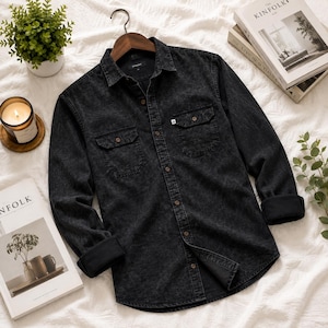 May include: A long-sleeve button-down shirt in a dark charcoal acid-wash denim fabric. The shirt features a classic collar, two chest pockets with buttoned flaps, and dark buttons. The cuffs are folded back to reveal a solid black interior lining.