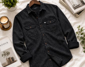 May include: A long-sleeve button-down shirt in a dark charcoal acid-wash denim fabric. The shirt features a classic collar, two chest pockets with buttoned flaps, and dark buttons. The cuffs are folded back to reveal a solid black interior lining.