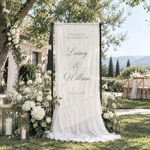 May include: A tall, off-white fabric wedding welcome sign suspended on a black metal frame. The text reads: WELCOME TO THE WEDDING OF Lainey & William JULY 15, 2026. The fabric features a diagonal ruffled layer across the lower section.