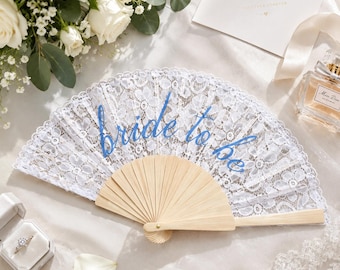 May include: A white lace hand fan with a light wooden frame and handle. The text "bride to be" is printed in blue cursive script across the centre of the lace. The fan is open and displays a detailed floral pattern throughout the white fabric.