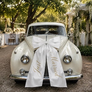 May include: A vintage cream-colored car decorated with a large white satin bow on the front hood. The bow features two long fabric tails hanging down the grille, with the gold cursive text "Just" on the left tail and "Married" on the right tail.
