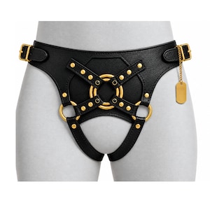 Könnte beinhalten: Ein schwarzer Kunstleder-Harness mit goldfarbenen Metallbeschlägen, bestehend aus kreisförmigen Ringen, Nieten und verstellbaren Seitenschnallen. Ein kleiner, rechteckiger goldfarbener Anhänger hängt an einer feinen Kette an der rechten Seite.