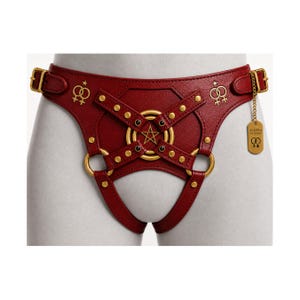 May include: A deep red textured harness featuring gold-toned hardware, including buckles, studs, and rings. The center displays a pentagram within concentric circles. Two female gender symbols are embossed on the sides. A gold tag reads AURORA FOR WOMEN.