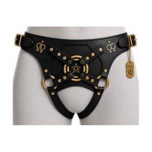 May include: A black harness featuring gold-toned hardware, including studs, rings, and a central pentagram emblem. The piece displays gold symbols of two interlocking female signs and a hanging tag with the text AURORA FOR WOMEN.