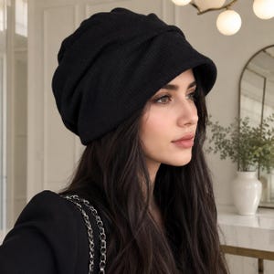 Könnte beinhalten: Eine einfarbige schwarze Beanie-Mütze mit einer weichen, locker sitzenden Silhouette und einem breiten, umgeschlagenen Rand. Der Stoff weist eine feine Rippstrickstruktur auf, die für ein strukturiertes und zugleich entspanntes Aussehen sorgt.