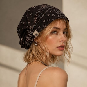 Könnte beinhalten: Ein schwarzes Kopftuch mit weißem Bandana-Muster aus geometrischen Formen und floralen Motiven. Der Stoff ist locker über den Kopf drapiert und verfügt über ein kleines, rechteckiges weißes Stoffetikett an der Seite mit einem dunklen Knopf.