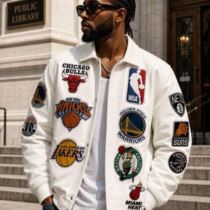 Puede incluir: Una chaqueta universitaria blanca con varios logotipos bordados de equipos de la NBA y el emblema oficial de la NBA. La chaqueta tiene cuello, cierre de botones a presión y puños acanalados. Se lleva sobre una camiseta blanca y vaqueros azules.