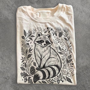 Puede incluir: Una camiseta de color crema con una ilustración en tinta negra de un mapache centrada en la parte delantera. El mapache está rodeado por un marco botánico detallado que consiste en varias hojas, piñas, bellotas y pequeñas bayas.
