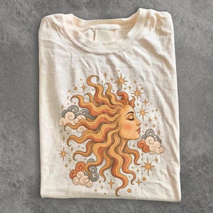 Può includere: Una t-shirt a maniche corte color crema con un'illustrazione grafica di un sole con volto umano, capelli ondulati e occhi chiusi. Il disegno è circondato da piccole stelle e nuvole stilizzate nei toni dell'arancione, del giallo e del grigio.