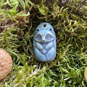 Pode incluir: Um pequeno pingente de formato oval com um guaxinim esculpido com acabamento azul perolado. O guaxinim é retratado com os olhos fechados e as patas cruzadas sobre o peito. Um pequeno orifício está posicionado no topo para fixação.