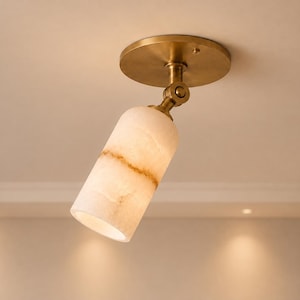 Può includere: Un faretto da soffitto con paralume cilindrico in pietra bianca traslucida con venature beige naturali. L'apparecchio include una base circolare in ottone e uno snodo regolabile, che consente di orientare la luce secondo le necessità.
