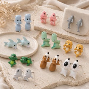 Peut inclure: Une collection de boucles d'oreilles à tige représentant des figurines d'animaux miniatures, notamment des requins, des dinosaures, des grenouilles et d'autres créatures aux couleurs variées comme le bleu, le vert, le rose, le jaune, le brun et le blanc.