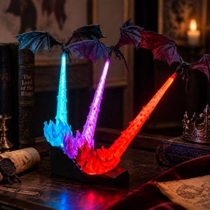 May include: A decorative lamp featuring three winged dragon figures, each emitting a vibrant beam of light in blue, purple, and red. The dragons are positioned above a shared black base, with each light beam rising from a textured, flame-like structure.