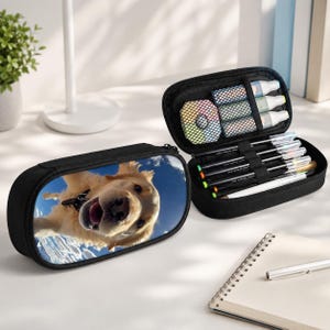 May include: A black zippered pencil case featuring a wide-angle photograph of a golden dog against a blue sky. The open case reveals an interior mesh pocket containing small round containers and elastic loops holding several pens and markers.