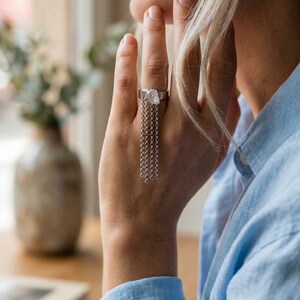 Puede incluir: Un anillo de plata con un engaste de metal texturizado e irregular que sostiene un grupo de delicadas cadenas de plata colgantes. Las cadenas cuelgan verticalmente de la banda, creando un efecto de cascada a lo largo del dedo.