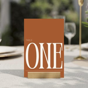 以下が含まれることがあります： バーントオレンジの背景に大きな白いセリフ体で「TABLE ONE」と書かれた長方形のテーブルナンバーカード。カードは長方形のゴールドトーンの金属製スタンドで直立しており、すっきりと整った印象を与えるイベント用の座席表示です。