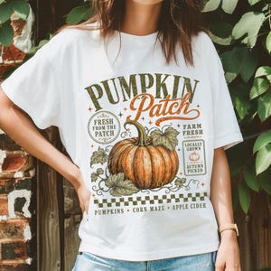 May include: A white t-shirt featuring a graphic of an orange pumpkin with green leaves. The text reads PUMPKIN Patch, FRESH FROM THE PATCH, FARM FRESH, LOCALLY GROWN, AUTUMN PICKED, PUMPKINS, CORN MAZE, and APPLE CIDER with a checkered border pattern.