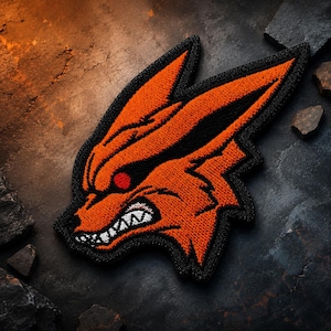 May include: An embroidered patch featuring the stylised head of a fox with bright orange fur, sharp black outlines, a single red eye, and a bared mouth showing white teeth. The design has a pointed ear and jagged fur details in a bold graphic style.