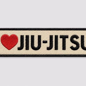 Könnte beinhalten: Ein rechteckiger gestickter Aufnäher mit beigem Hintergrund und schwarzem Rand. Der Text lautet I, gefolgt von einem roten Herzsymbol und dem Wort JIU-JITSU in fetten schwarzen Großbuchstaben. Das Design ist auf hellgrauem Grund zentriert.