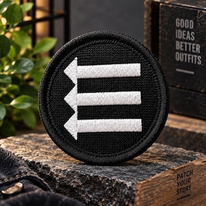 May include: A circular black embroidered patch featuring a white graphic design of three horizontal arrows pointing to the left, stacked vertically. The patch has a thick black stitched border and a textured fabric surface with high-contrast white thread.