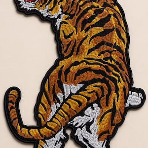 Op de afbeelding: Een geborduurde patch met een tijger in een dynamische houding met de bek open. De tijger heeft een oranje vacht met zwarte strepen, witte accenten op het gezicht, de borst en de poten, en een rode tong. Het ontwerp is omlijnd met zwart garen.