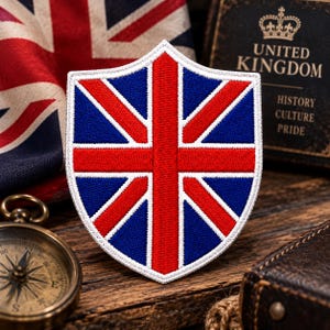 May include: A shield-shaped embroidered patch featuring the Union Jack flag design in red, white, and blue. The patch has a distinct white border and displays the iconic cross and diagonal stripes of the United Kingdom flag in a structured, stitched fabric format.