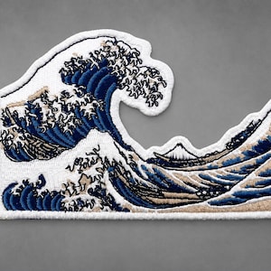 May include: An embroidered patch featuring a detailed depiction of a large crashing ocean wave in shades of dark blue, white, and tan. The design includes a small mountain peak in the background, all outlined with a thick white stitched border.