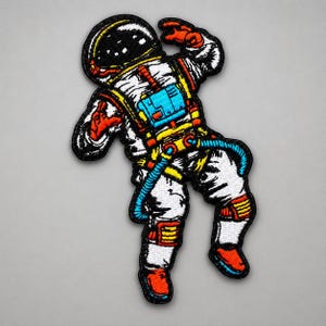 May include: An embroidered patch featuring an astronaut in a white spacesuit with black, orange, yellow, and blue accents. The astronaut is depicted in a floating pose with a black helmet visor, orange gloves, and orange boots against a plain background.