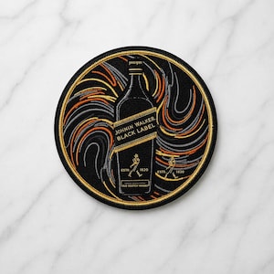 May include: A circular embroidered patch featuring a black Johnnie Walker Black Label whisky bottle silhouette in the center. The design includes gold, orange, and grey swirling lines on a black background with the text Johnnie Walker Black Label.
