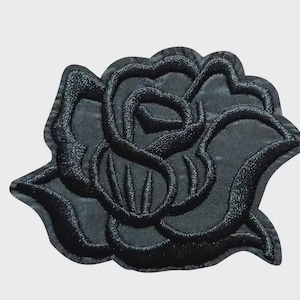 以下が含まれることがあります： 黒い刺繍のバラのワッペン。密度の高い糸で立体的な花の質感を表現しています。花びらは太いステッチの輪郭で縁取られており、重なり合うバラの形を形成しています。全体的にマットな黒色で仕上げられています。