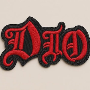 Puede incluir: Un parche bordado que presenta la palabra DIO en una fuente roja estilizada de inspiración gótica con un borde negro grueso. Las letras están cosidas con una textura de hilo densa, creando una apariencia en relieve sobre el fondo de tela negra.