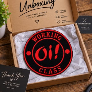 Könnte beinhalten: Ein kreisförmiger gestickter Aufnäher mit schwarzem Hintergrund und rotem Rand. Der Text "WORKING" ist oben gebogen, "CLASS" unten gebogen und "Oi!" ist mittig in Rot platziert. Der Aufnäher liegt in einer Pappschachtel mit Seidenpapier.
