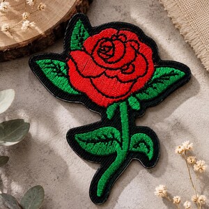 May include: An embroidered patch featuring a vibrant red rose with green leaves and a green stem, all outlined in black thread. The design is set against a black background, creating a clear contrast between the floral elements and the dark border.