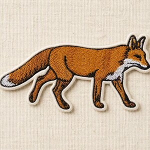Könnte beinhalten: Ein gestickter Aufnäher in Form eines laufenden Fuchses. Der Fuchs hat einen rostorangen Körper mit schwarzen Akzenten an den Beinen und Ohren sowie einen weißen Fleck an Brust, Gesicht und Schwanzspitze. Das Design hat einen cremefarbenen Rand.