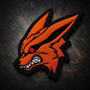 May include: An embroidered patch featuring a stylised orange fox head with a black border. The design displays a snarling expression with visible white teeth, a single red eye, and pointed ears, rendered in dense orange thread on a black background.