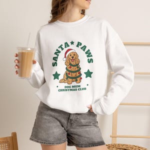 May include: A white long-sleeve sweatshirt featuring a graphic of a golden dog wearing a Santa hat and wrapped in a garland with ornaments. The text reads SANTA PAWS and DOG MUM CHRISTMAS CLUB, accompanied by green stars and small dots.