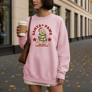 May include: A light pink crewneck sweatshirt featuring a graphic of a golden dog wearing a Santa hat and wrapped in Christmas lights. The text reads SANTA PAWS and DOG MUM CHRISTMAS CLUB, accompanied by red stars on a light pink fabric background.