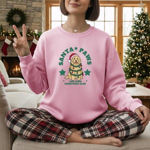 May include: A light pink crewneck sweatshirt featuring a graphic of a golden-colored dog wearing a Santa hat and wrapped in Christmas lights. The text reads SANTA PAWS and DOG MUM CHRISTMAS CLUB, surrounded by green stars on the front of the garment.