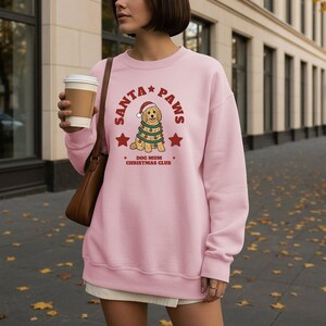 May include: A light pink crewneck sweatshirt featuring a graphic of a golden retriever wearing a Santa hat and wrapped in Christmas lights. The text reads SANTA PAWS and DOG MUM CHRISTMAS CLUB in red lettering, accented by two red stars on the sides.