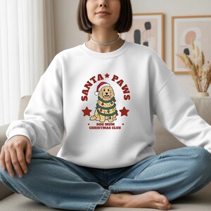May include: A white crewneck sweatshirt featuring a graphic of a golden-colored dog wearing a Santa hat and wrapped in a string of colorful Christmas lights. The text reads SANTA PAWS, DOG MUM CHRISTMAS CLUB, accompanied by two red stars.