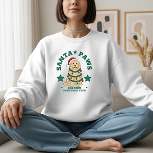 May include: A white long-sleeve sweatshirt featuring a graphic of a golden-colored dog wearing a Santa hat and wrapped in colorful Christmas lights. The text reads SANTA PAWS and DOG MUM CHRISTMAS CLUB in green lettering with two green stars.