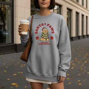 May include: A heather gray crewneck sweatshirt featuring a graphic of a golden dog wearing a Santa hat and wrapped in Christmas lights. The text reads SANTA PAWS and DOG MUM CHRISTMAS CLUB in red, accompanied by red star icons on the front chest.