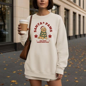 May include: A cream-colored crewneck sweatshirt featuring a graphic of a golden-colored dog wearing a Santa hat and wrapped in Christmas lights. The text reads SANTA PAWS and DOG MUM CHRISTMAS CLUB, accented by two red stars on the front.