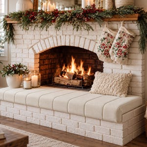 May include: A white brick fireplace hearth features a cream-colored upholstered bench cushion, a textured throw pillow, two floral patterned Christmas stockings, and a festive garland with red berries and greenery above the burning fire.